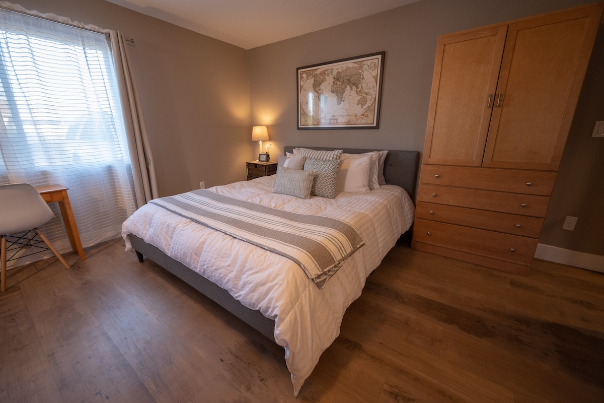 A cozy sleeping area features a spacious bed dressed in neutral linens, accompanied by a tufted headboard. A dresser stands next to a window adorned with sheer curtains, allowing soft light to illuminate the room. A comfortable chair is positioned against the wall.