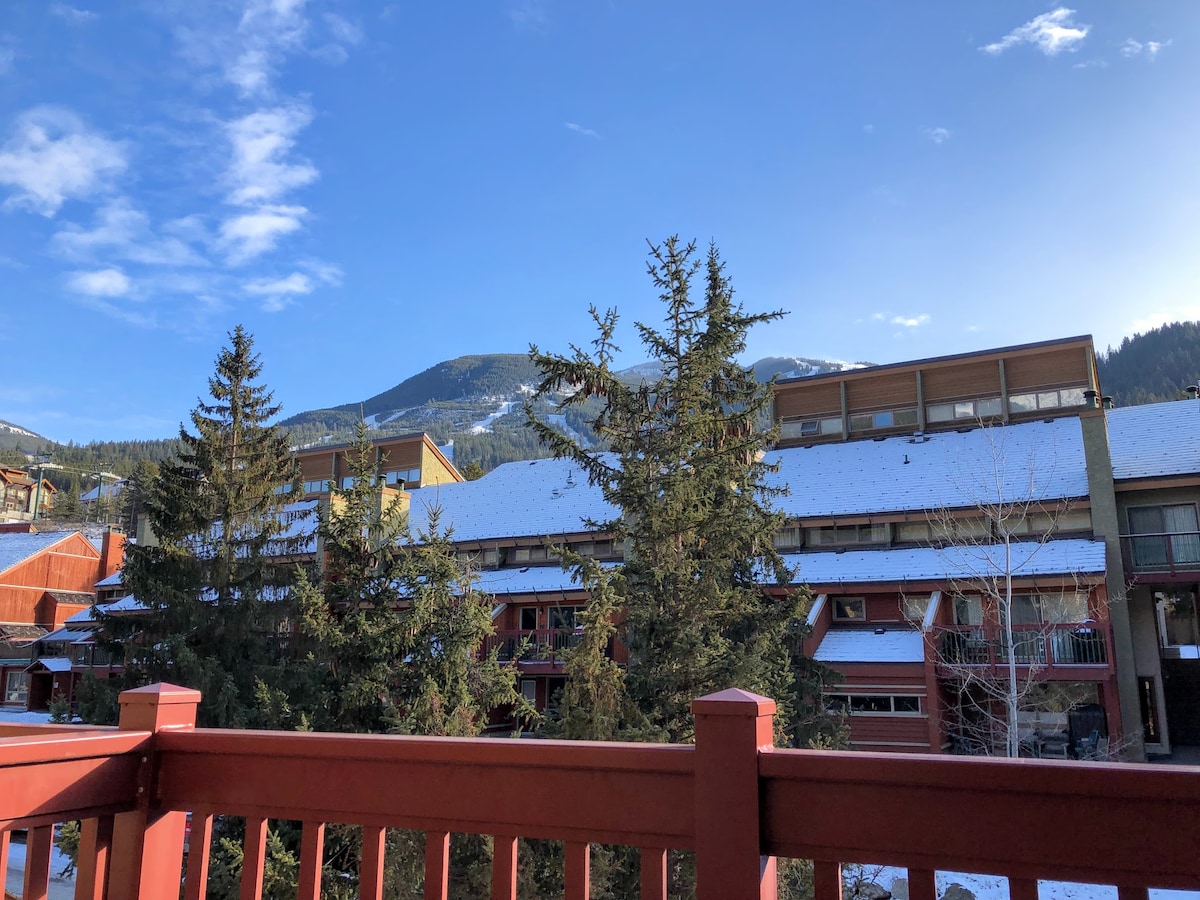 A view from the patio showcases surrounding mountains and evergreen trees. The sky is clear, and snow covers some rooftops, hinting at a winter atmosphere. The railing of the patio is made of wood, providing a warm contrast to the natural landscape.