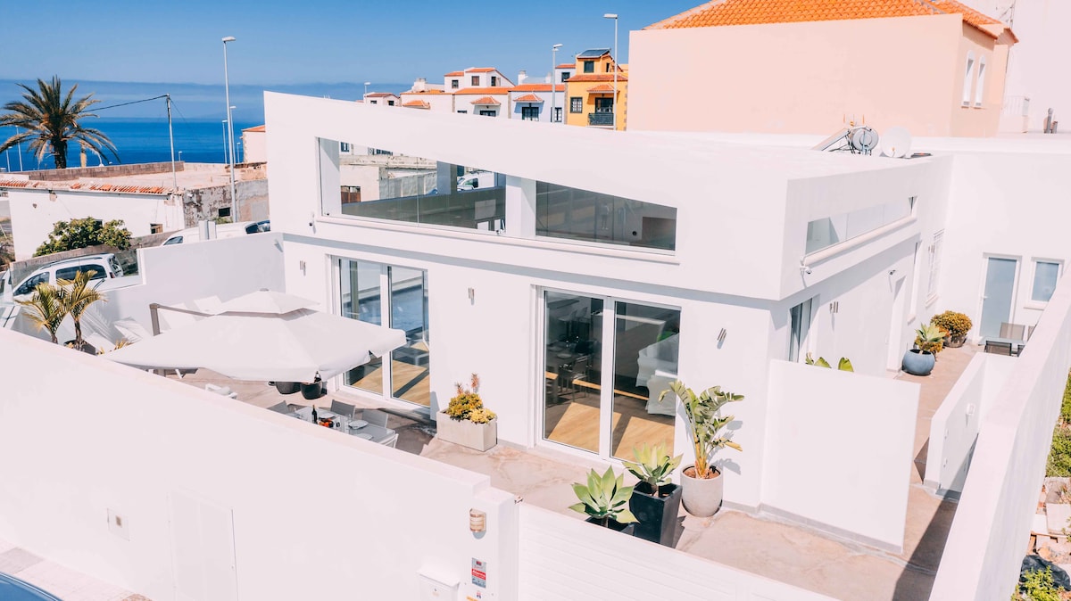 The exterior view of the villa showcases a modern architectural design, highlighted by large glass doors and white walls. An outdoor seating area is presented alongside a shaded umbrella and several potted plants, with a clear view of the surrounding residential area and ocean in the background.