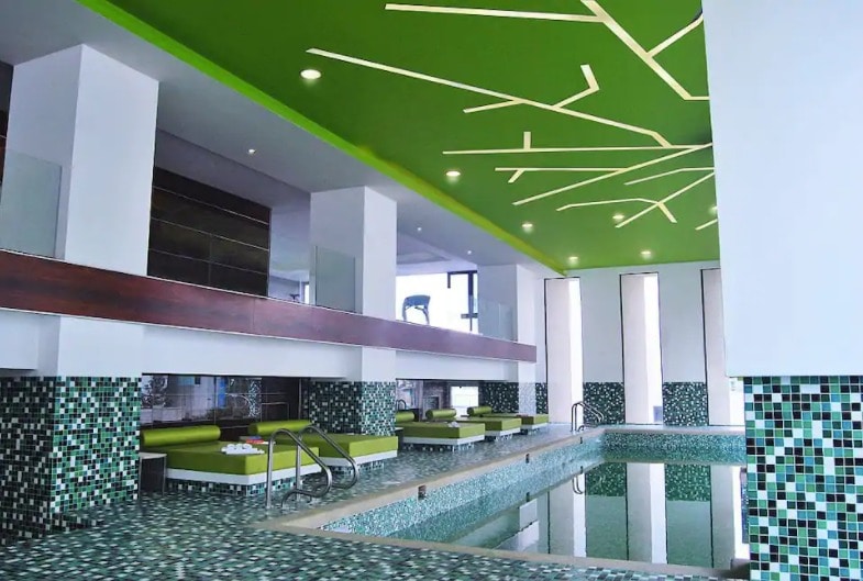 An indoor pool area features a modern design with vibrant green ceiling accents. Green lounge chairs are positioned along the edges of the pool, which is surrounded by decorative tile work in shades of blue and green. Large windows provide natural light.