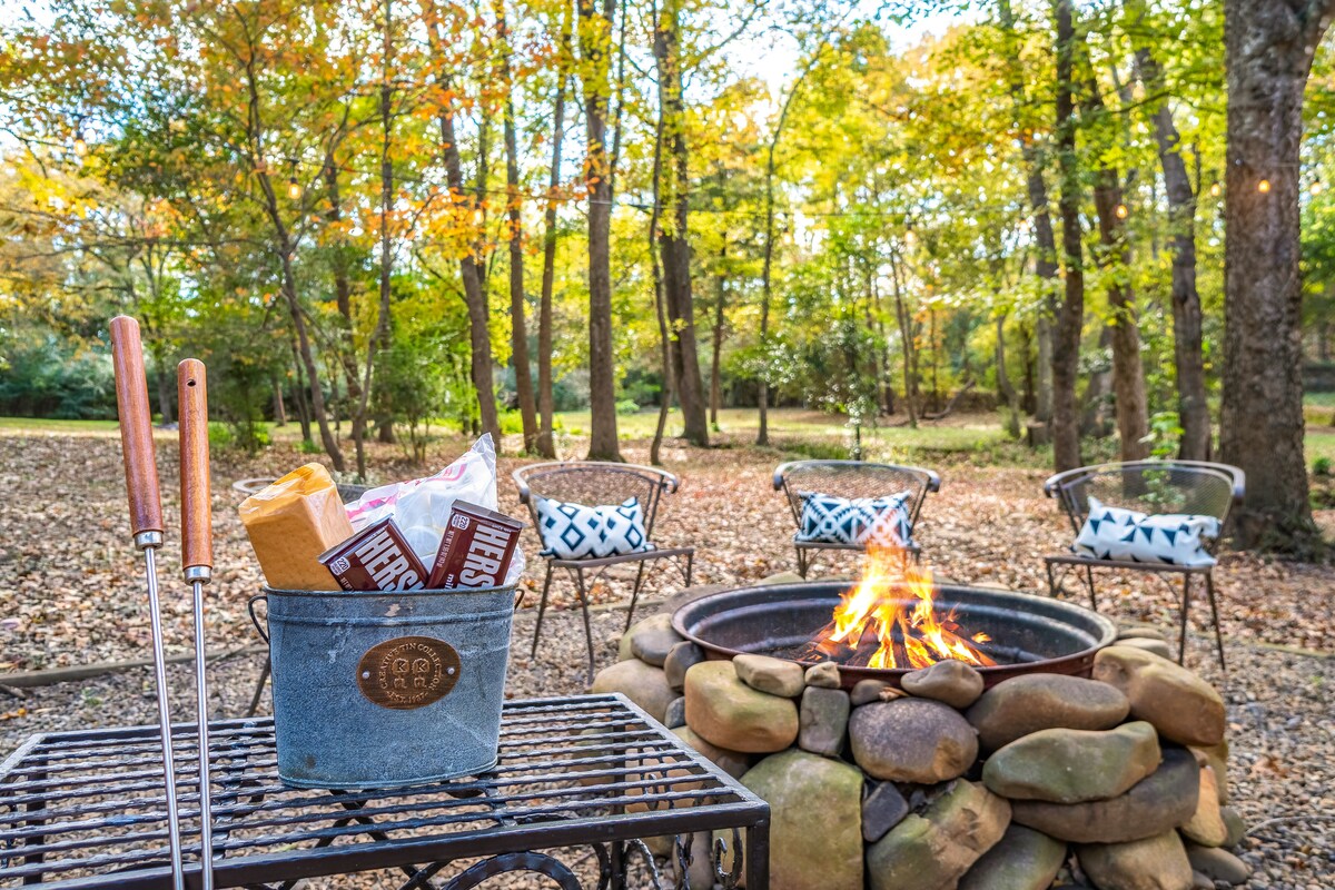 An inviting fire pit area is surrounded by natural stones, with a warm fire crackling in the center. Four chairs are positioned nearby, featuring patterned cushions. A tin bucket holding s'mores ingredients is placed on a wicker table, accentuating the outdoor atmosphere amidst lush trees.