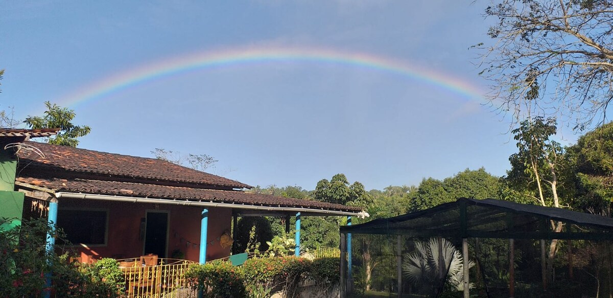 A rainbow stretches across a clear sky above a charming countryside house. The house is surrounded by lush greenery and trees, creating a serene landscape. A netted area is visible, adding to the natural ambiance of the setting.