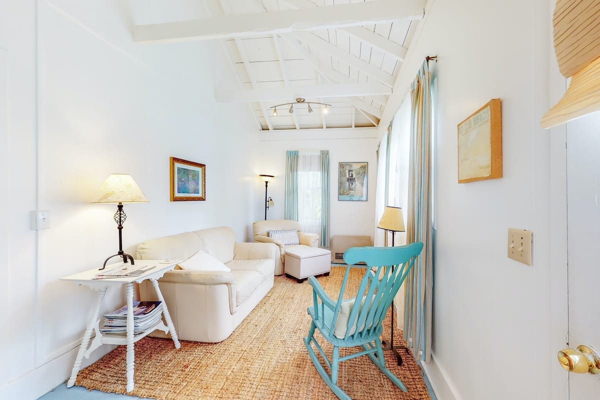 A light-filled living area features a cozy white sofa, an armchair, and a rocking chair, arranged around a small table. Natural light enters through large windows, with soft blue curtains framing the scene. A woven rug covers the floor, adding texture to the space.