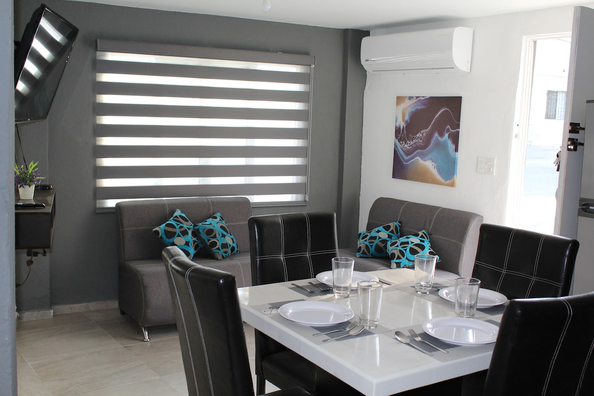 An open dining area is paired with a cozy seating space. A table set for four features clear plates and glasses. A grey sofa with decorative blue cushions complements the contemporary decor, while a mounted television adds convenience. Natural light filters through the shades.