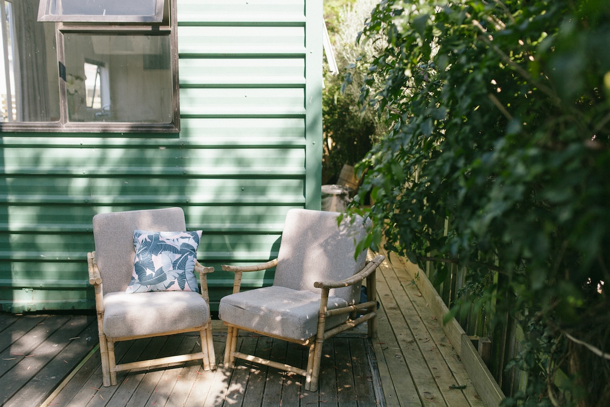 Two chairs made of natural wood are situated on a wooden deck beside a green cabin. A decorative pillow with a floral pattern rests on one chair. Dense greenery surrounds the area, providing a sense of privacy and tranquility.