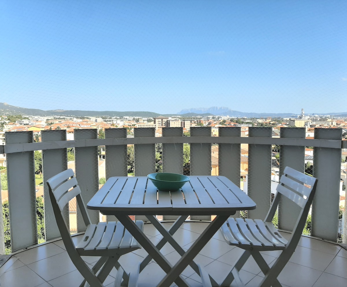 A panoramic veranda is depicted, featuring a small white table with two matching chairs. The surrounding railing offers an unobstructed view of the city and distant mountains, with clear blue skies enhancing the scene.