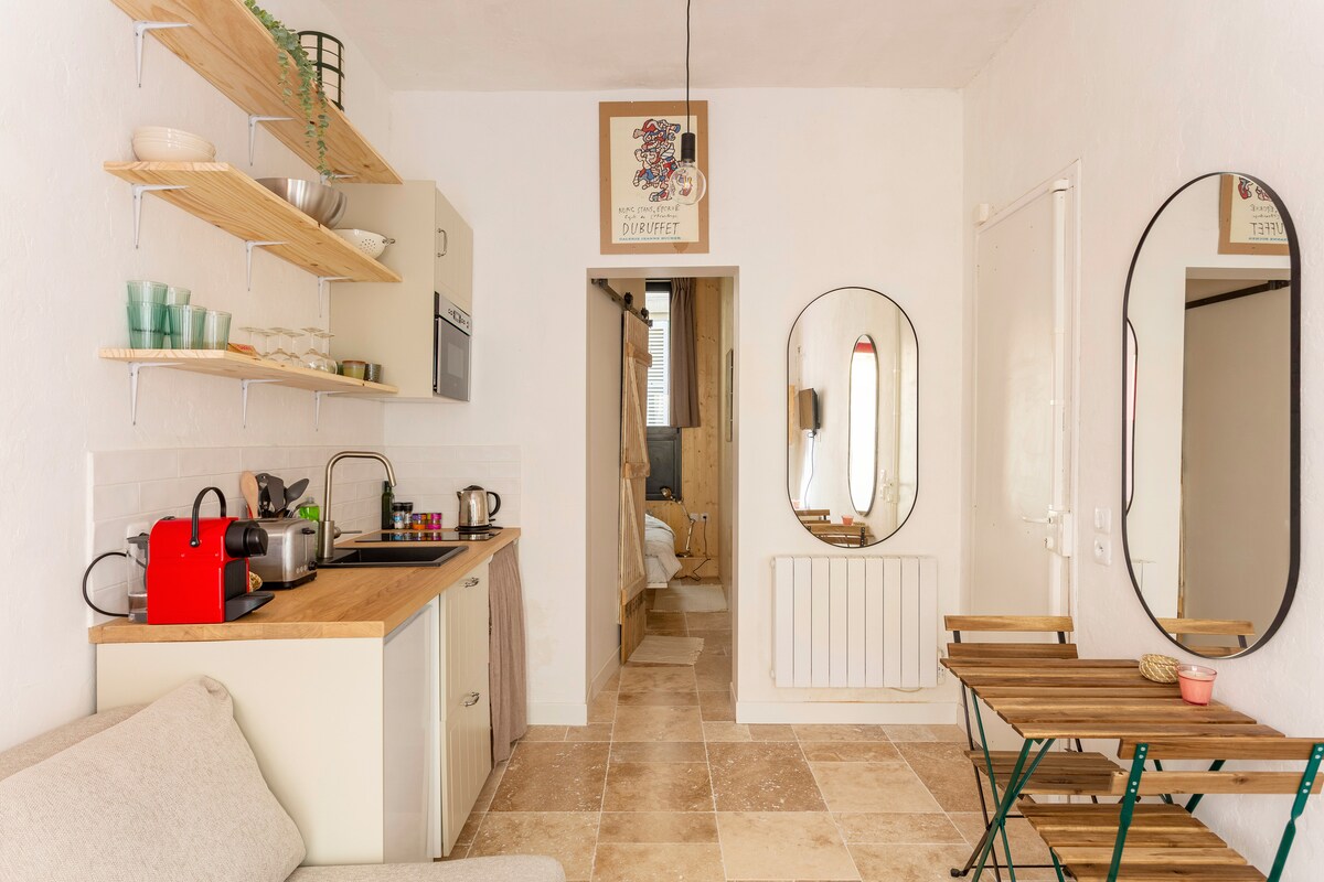 A compact kitchen area is outfitted with essential appliances and minimalist shelving. A small dining table with two chairs is positioned near a large mirror, reflecting the cozy interior. Neutral tones and natural light contribute to a warm ambiance.