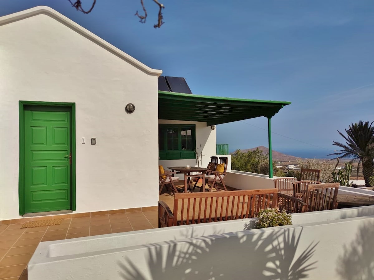 A white building with a distinctive green door is depicted, featuring an outdoor terrace furnished with wooden chairs and a table under a green awning. Views of the surrounding landscape and ocean can be seen in the background, enhancing the serene setting.