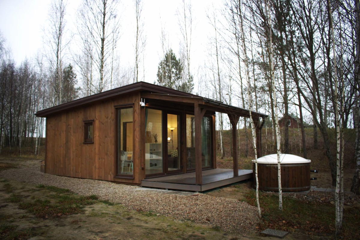 A wooden cabin is surrounded by bare trees, featuring large glass doors that invite natural light. A covered terrace provides a seating area, while a hot tub is positioned nearby, offering a tranquil outdoor space for relaxation.