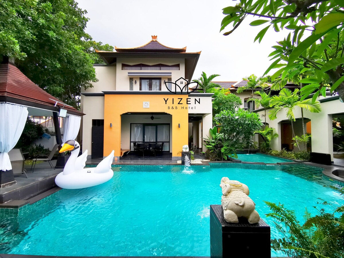 A vibrant outdoor scene features a tropical pool surrounded by lush greenery. A large, inflatable swan floats in the water, while artistic sculptures enhance the tranquil atmosphere. The villa's classic architecture is complemented by a spacious terrace, inviting relaxation in the serene environment.
