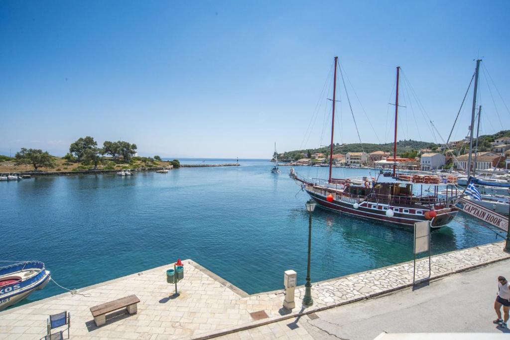 A calm waterfront scene displays boats gently moored at a serene harbor. The clear blue water reflects the cloudless sky, and lush greenery frames the background. A stone pathway leads towards the water's edge, complemented by nearby benches and light posts.