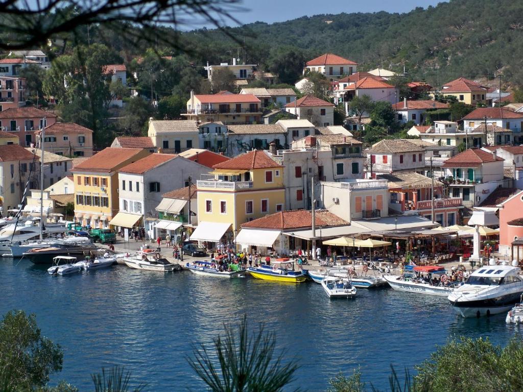 The image captures a bustling seaside village with colorful buildings lining a picturesque waterfront. Numerous boats are docked in the calm blue water, and several outdoor dining areas are filled with people enjoying their meals. Lush greenery frames the scene, complementing the relaxed atmosphere.