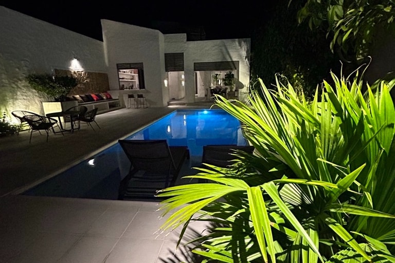 A serene nighttime view of a swimming pool is captured, with gentle illumination reflecting from the water. Lush green plants frame the pool area, while seating arrangements are discreetly placed nearby, creating an inviting outdoor space.