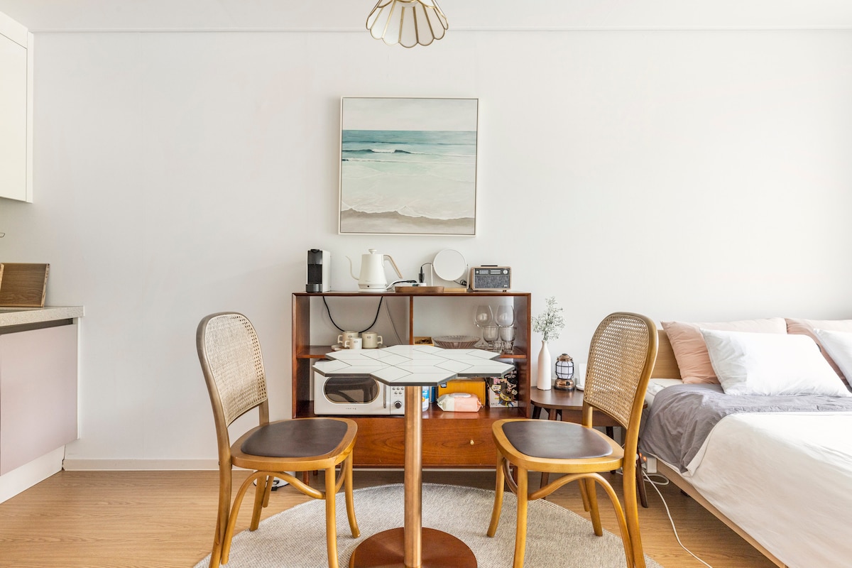 A small dining area features a round table surrounded by two wooden chairs, creating a cozy nook. A low wooden shelf holds kitchen essentials and glassware. A painting of a serene coastal scene adorns the wall, complementing the light color scheme of the room.