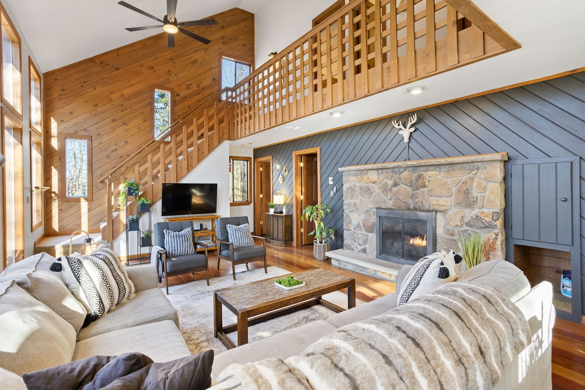 The spacious living area features a stone fireplace framed by comfortable seating, fostering an inviting environment. Sunlight streams through multiple large windows, highlighting the wood-paneled walls and wooden staircase. A television is mounted on the wall opposite the fireplace, enhancing the entertainment options.