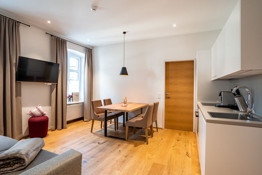 The living area features a modern dining table with four chairs adjacent to a fully equipped kitchenette. A small accent chair adds a pop of color. Natural light fills the space through a large window, complemented by soft, neutral-toned drapes. A mounted television is positioned on the wall.