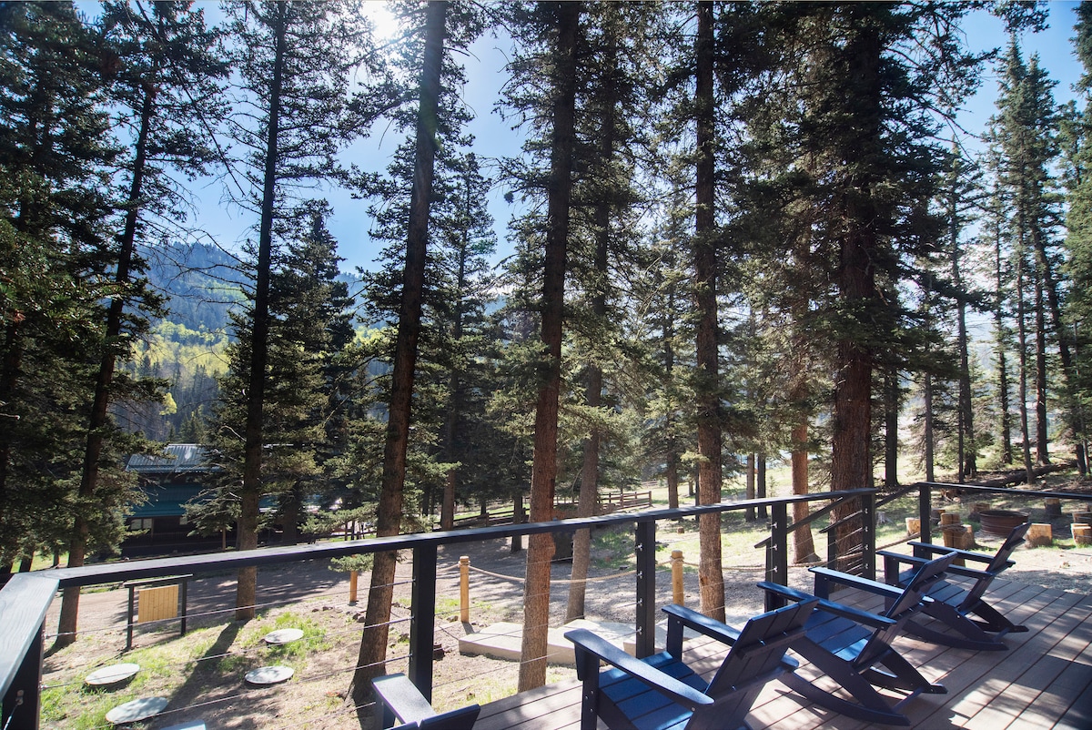 A deck features four Adirondack rocking chairs, providing a comfortable spot to enjoy the surrounding forest. Towering pine trees create a natural backdrop, and sunlight filters through the branches, enhancing the peaceful nature setting.