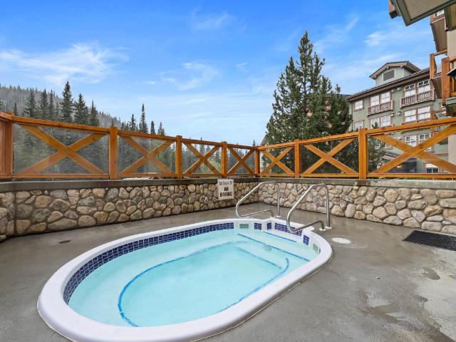 An outdoor hot tub is set against a backdrop of pine trees, featuring a curved shape and submerged steps for easy access. Brown wooden fencing surrounds the area, providing a sense of privacy while highlighting the scenic view of the surrounding landscape.