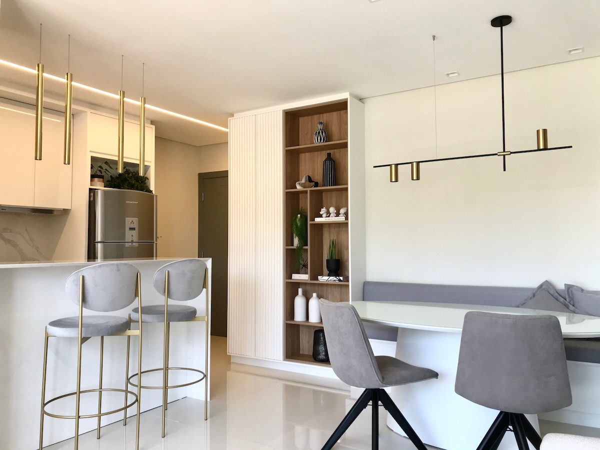 The modern kitchen features sleek cabinetry and stainless steel appliances, complemented by gold-accented bar stools. A dining area is highlighted by a minimalist chandelier above a light-colored table, with a built-in shelf displaying decorative items and a cozy seating nook.