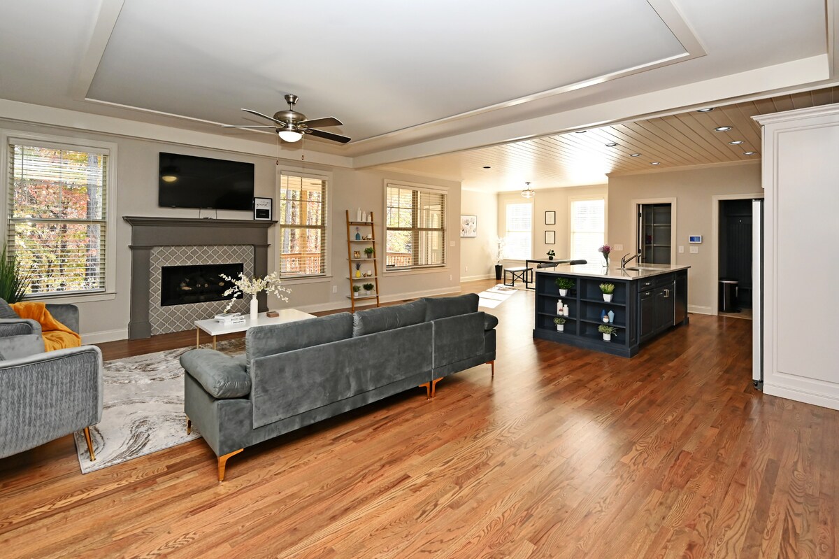 An open-concept living area is highlighted by hardwood floors and ample natural light. A modern gray sofa faces a decorative fireplace and television. An island with bar seating is visible in the background, complemented by a dining area bathed in sunlight from large windows.