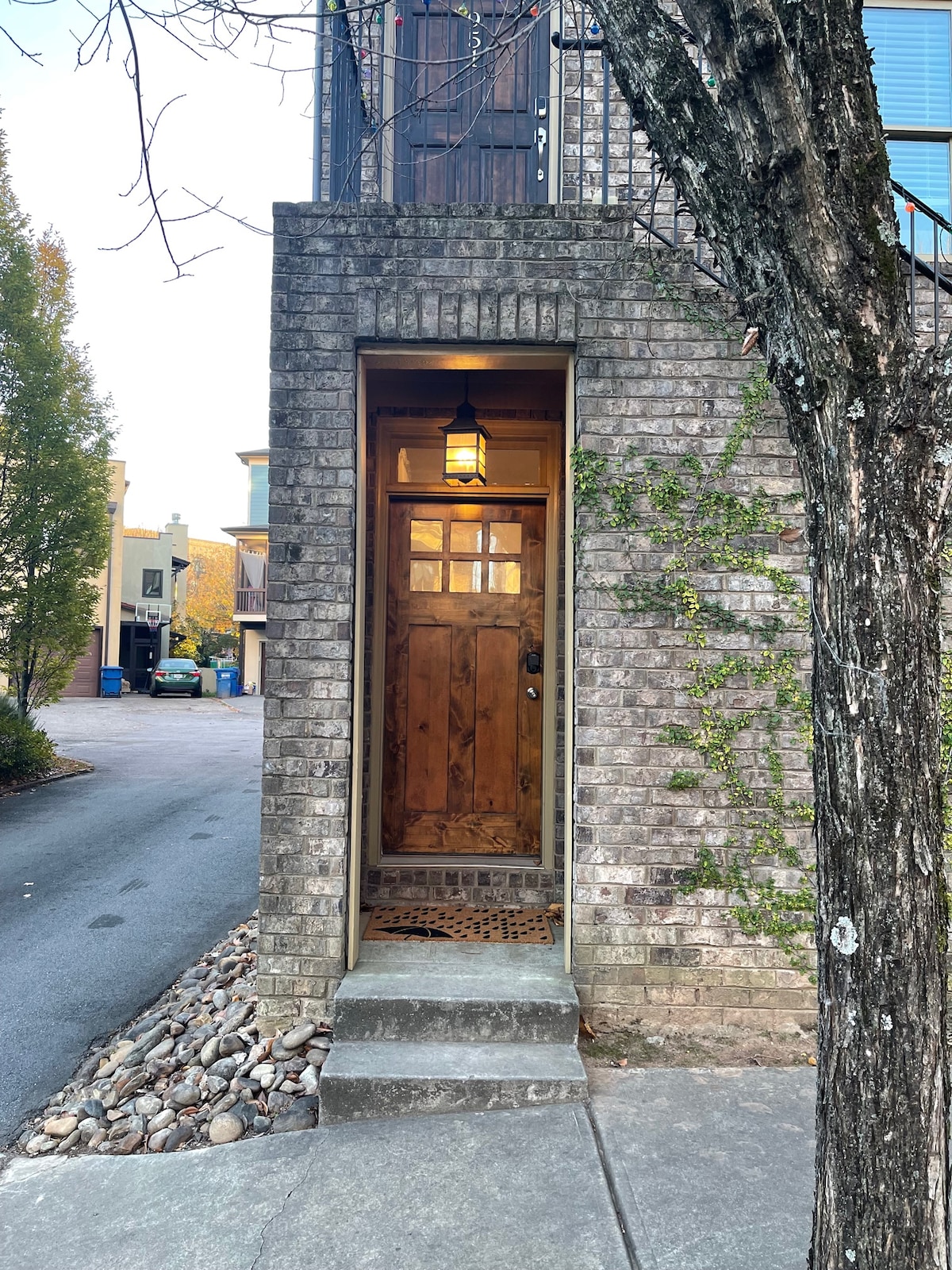 A sturdy wooden door with glass panels is framed by a brick facade, creating an inviting entrance. A small light fixture hangs above the door, illuminating the stone pathway lined with irregularly shaped pebbles. The surroundings include tree branches and hints of greenery.