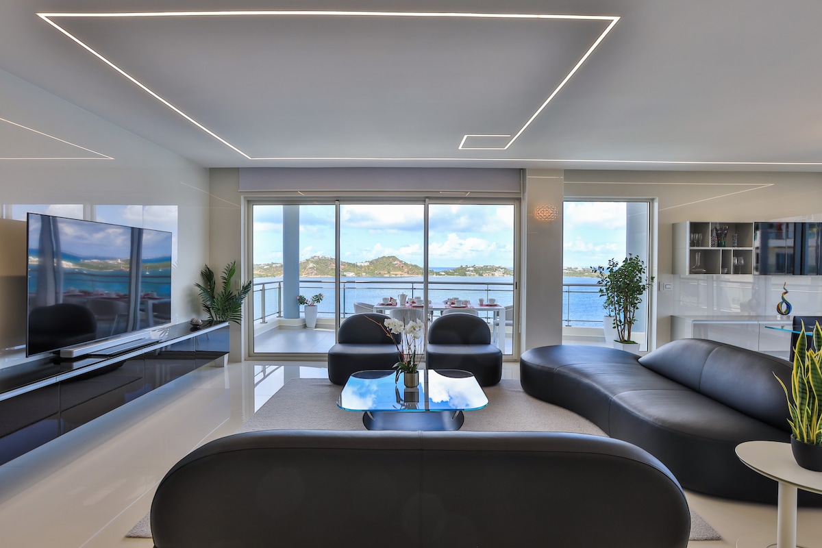 A modern living area features sleek black sofas arranged around a glass coffee table. Large windows provide panoramic views of the surrounding landscape and water, while soft, ambient lighting enhances the room's clean lines and sophisticated design.