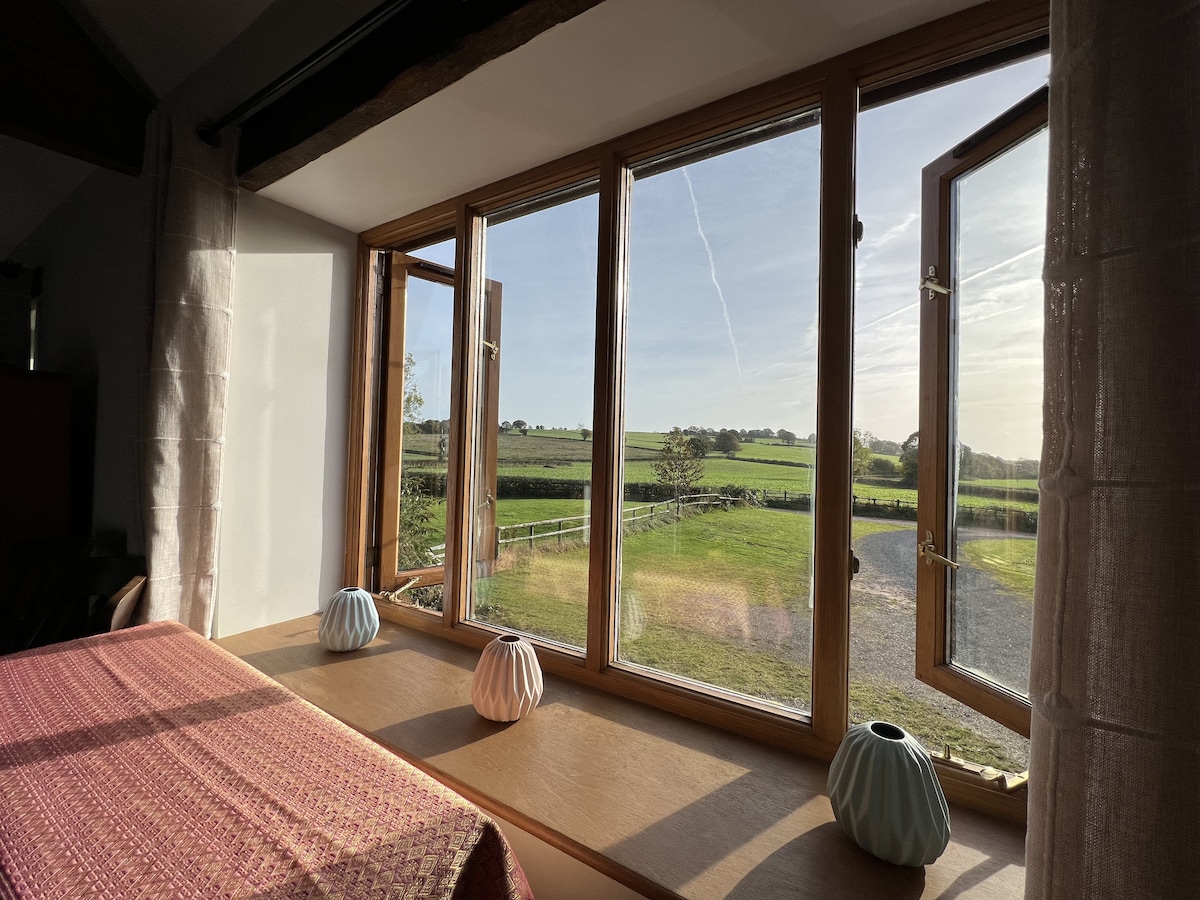 Large windows frame panoramic views of the open countryside, allowing natural light to fill the room. Decorative vases in soft colors are positioned on the windowsill. The tranquil setting showcases green fields and a clear sky, creating an inviting atmosphere.