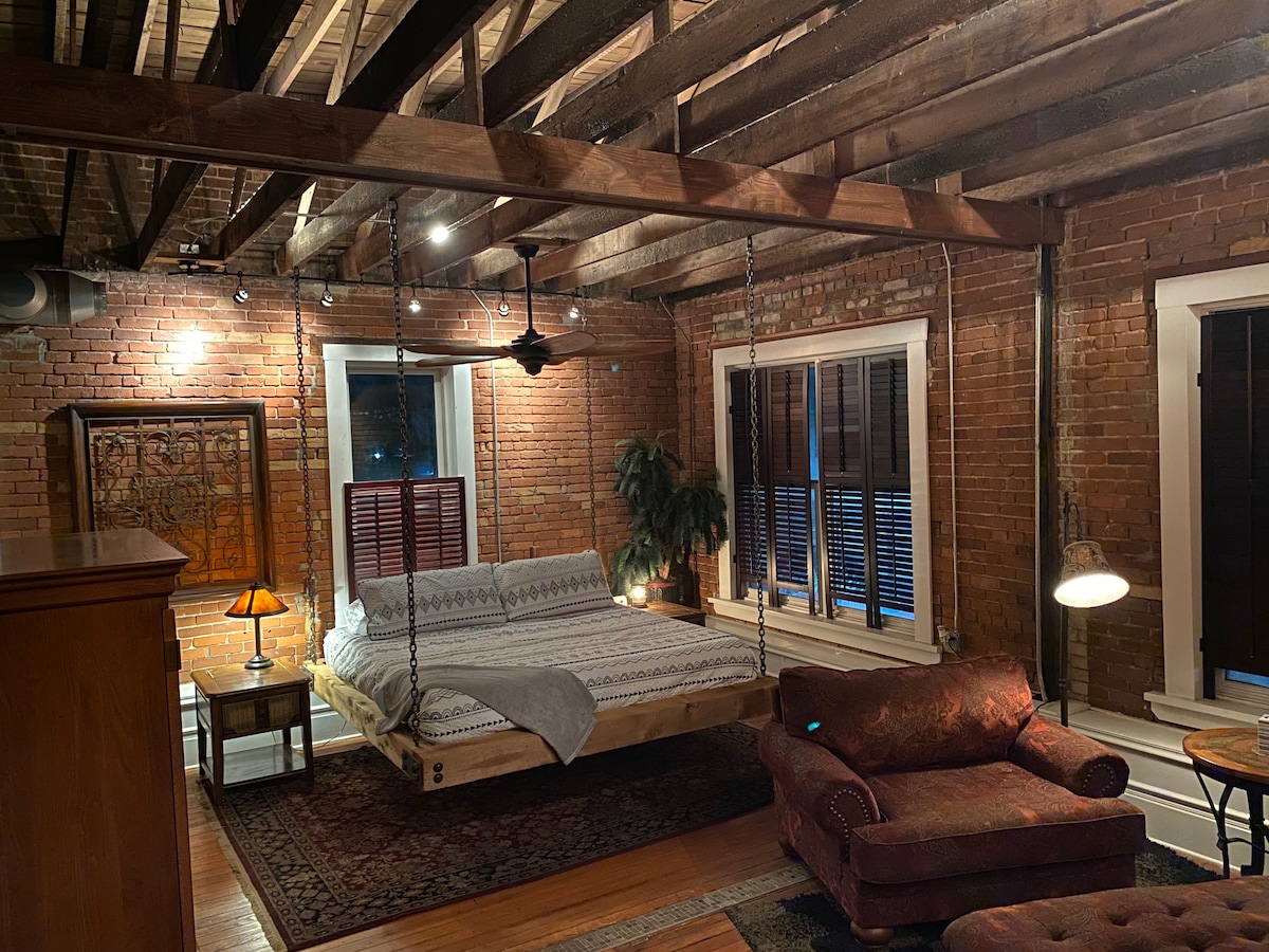 A spacious master bedroom features open rafters and a king-size bed suspended by log chains. Exposed brick walls add character, while a patterned blanket is draped over the bed. A plant and vintage lighting enhance the charming ambiance of the room.