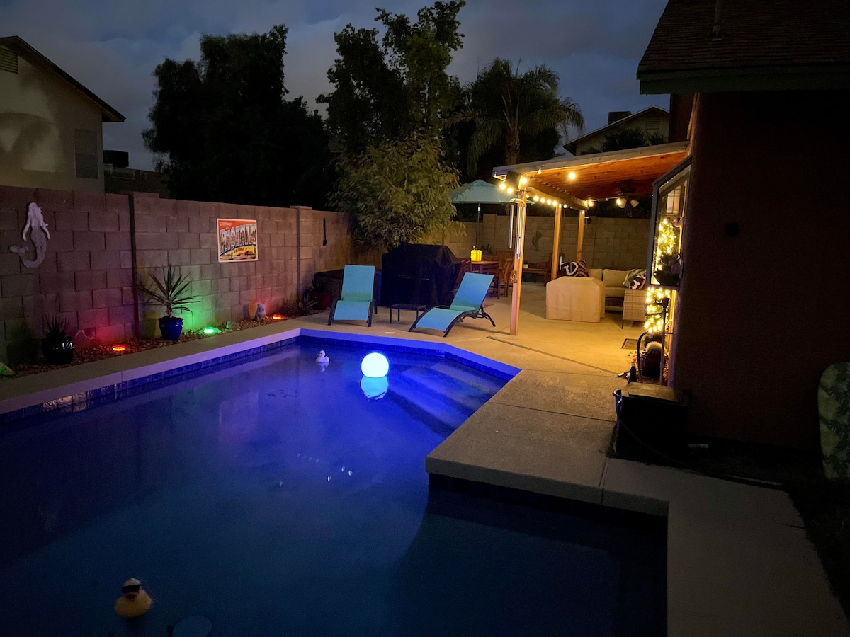 The pool area is softly illuminated by string lights, creating a relaxing ambiance. A colorful light floats in the water. Comfortable seating is set on the patio, surrounded by lush greenery, with a view of the tranquil pool, inviting evening enjoyment.