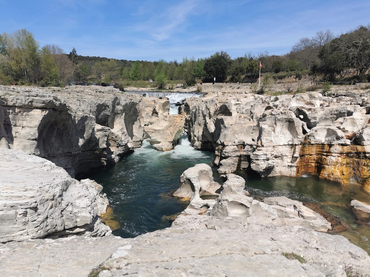 A rocky river landscape features smooth stone formations surrounding tranquil waters. The river flows gently through the crevices, reflecting the bright blue sky and greenery in the background. Trees line the banks, contributing to the serene atmosphere.