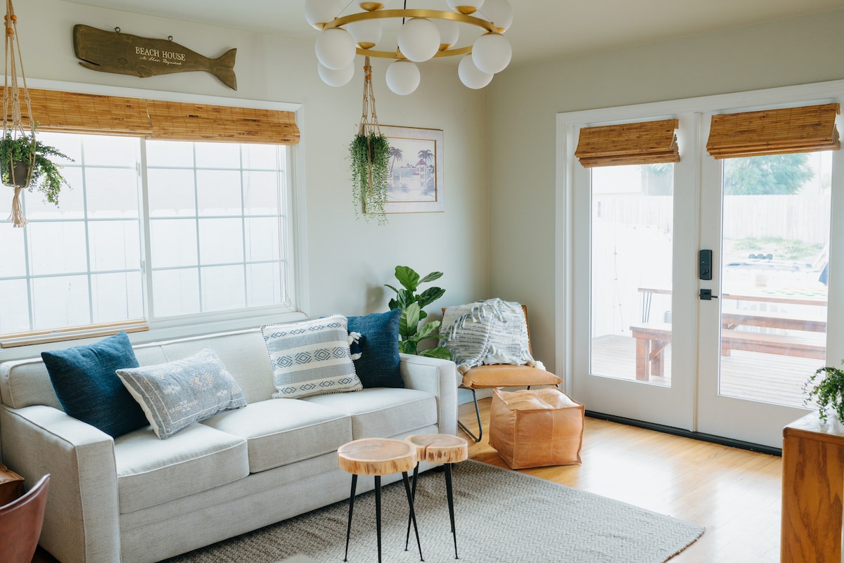 A bright living area is featured, showcasing a light-colored sofa adorned with decorative pillows. Two large windows allow natural light to fill the space, complemented by bamboo shades. A wooden side table and a potted plant add warmth, while French doors provide access to the deck.