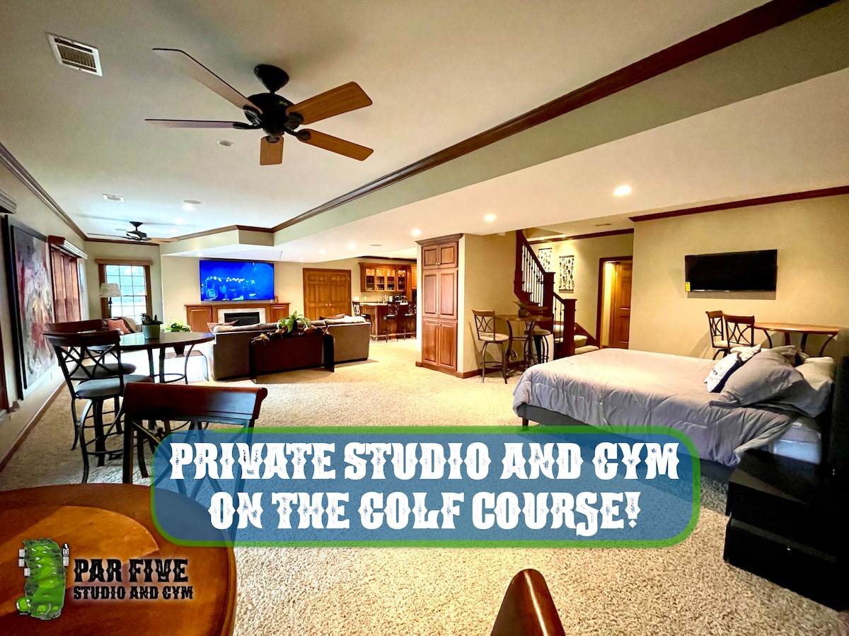 A spacious studio area features a king bed and an open living space with a dining area. Two ceiling fans provide comfort, and large windows allow natural light. A kitchenette is visible, alongside a desk with an ultrawide monitor, enhancing the room's versatility.