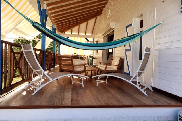 A covered porch features a relaxing hammock strung between wooden posts and several chairs arranged around a circular table. Bright awnings provide shade, while wooden flooring and white railings enhance the inviting outdoor space.