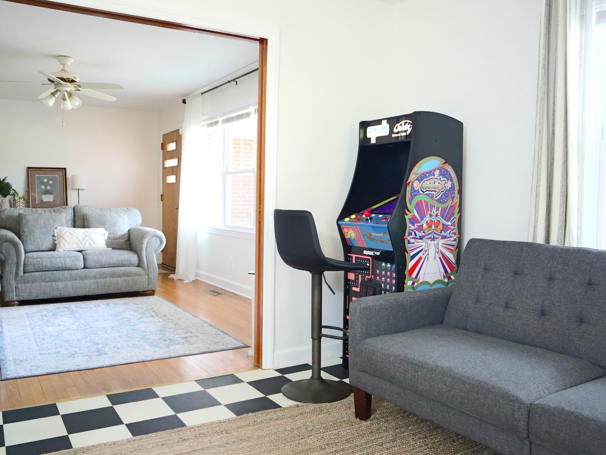 A cozy living area is divided by an open doorway, featuring a comfortable gray sofa and a stylish arcade machine. The flooring contrasts with a checkered pattern, while natural light floods in from nearby windows adorned with light curtains.