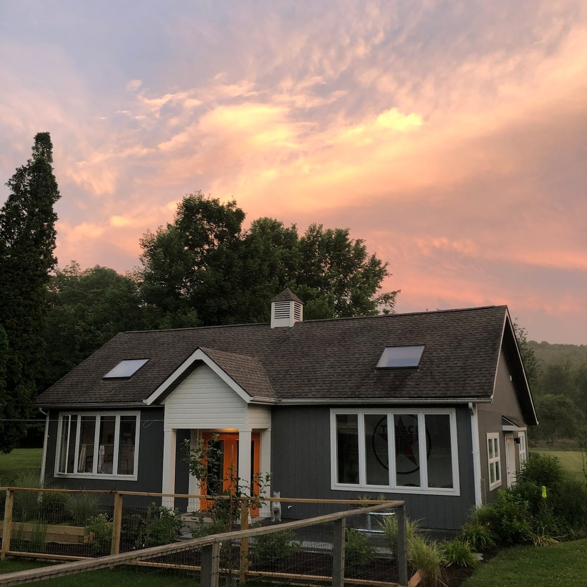 The exterior of a modern guest cottage is shown, with a gray facade and vibrant orange doors. Skylights are visible on the roof, framed by lush trees. A wooden fence surrounds the property, creating a welcoming outdoor space under a colorful sunset sky.