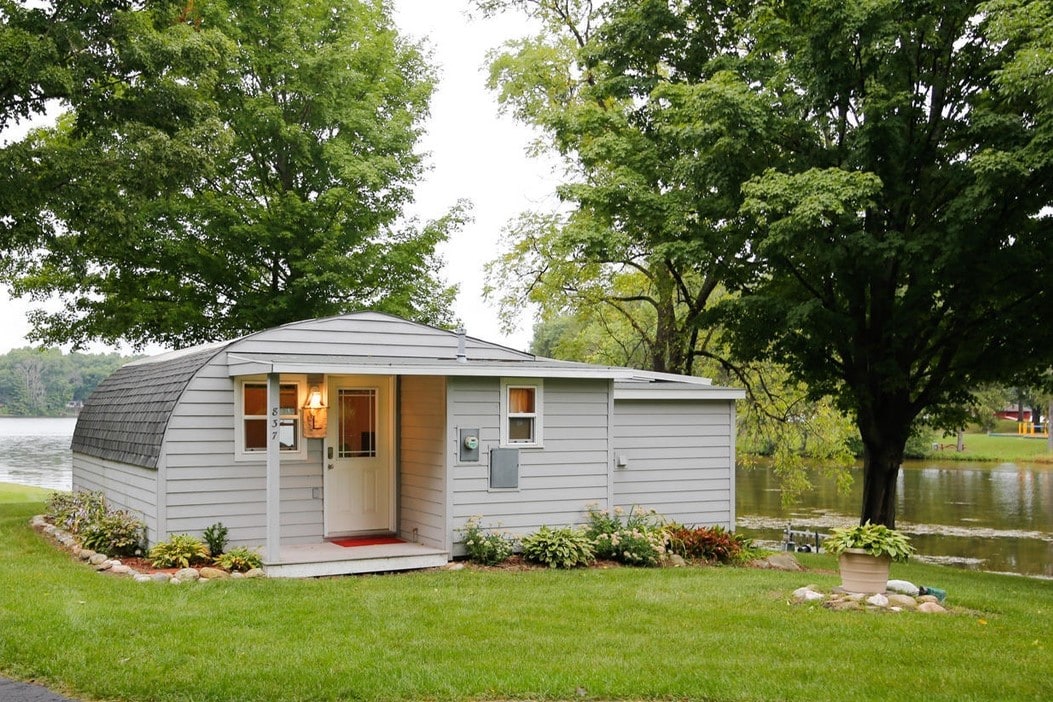 A charming Quonset hut is set against the backdrop of a serene lake. The structure features a welcoming entrance with a red accent rug, surrounded by a lush grassy yard and garden beds. Majestic trees provide shade and enhance the natural landscape.