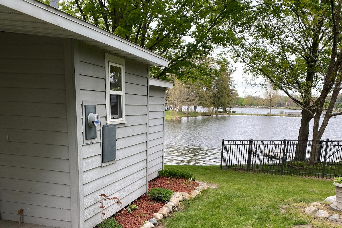 A side view of the Quonset Hut is showcased, featuring the gray wooden exterior and a small window. Lush green grass and garden stones lead up to a fenced area adjacent to the tranquil waters of Maple Lake, surrounded by trees.