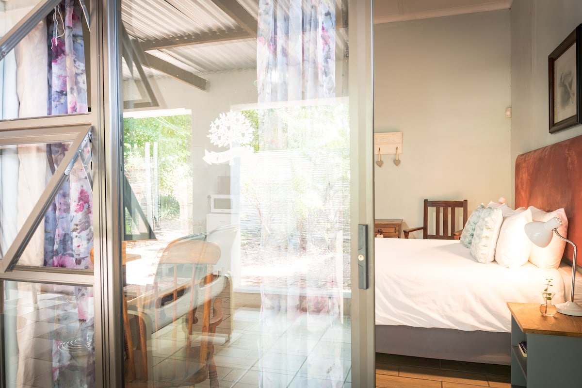 A serene bedroom is viewed through large glass doors, featuring a spacious bed with light-colored bedding. Bright floral curtains frame the windows, allowing natural light to fill the room. A wooden chair and a small table are positioned nearby, adding a cozy touch to the space.