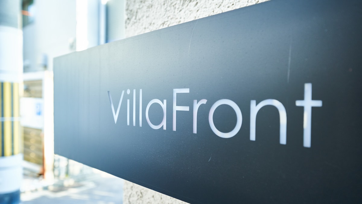 A close-up view of the VillaFront sign is displayed against a textured wall. The sign features modern, clear lettering, providing easy identification of the property. The background includes blurred elements, suggesting a welcoming entryway.