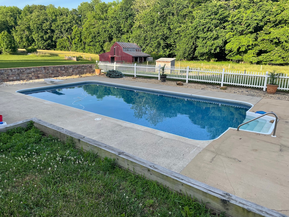 A private pool features a clear blue surface, surrounded by a concrete patio. Lush greenery borders the area, while a wooden railing encloses the space. In the background, a barn and a greenhouse complement the serene farm setting.