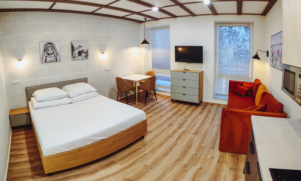 A spacious studio apartment features a queen-sized bed with crisp white linens. A flat-screen Smart TV is mounted on the wall, facing a small dining table with two chairs. An orange sofa adds a pop of color, while large windows allow natural light to enter.