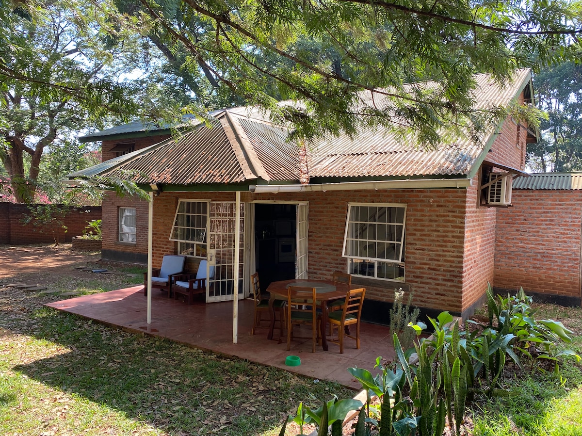 The exterior of a brick house features a covered porch with seating arrangements. A small wooden table and chairs are situated on the porch, surrounded by greenery. The roof has a gentle slope, and large windows enhance the bright and open appearance of the house.