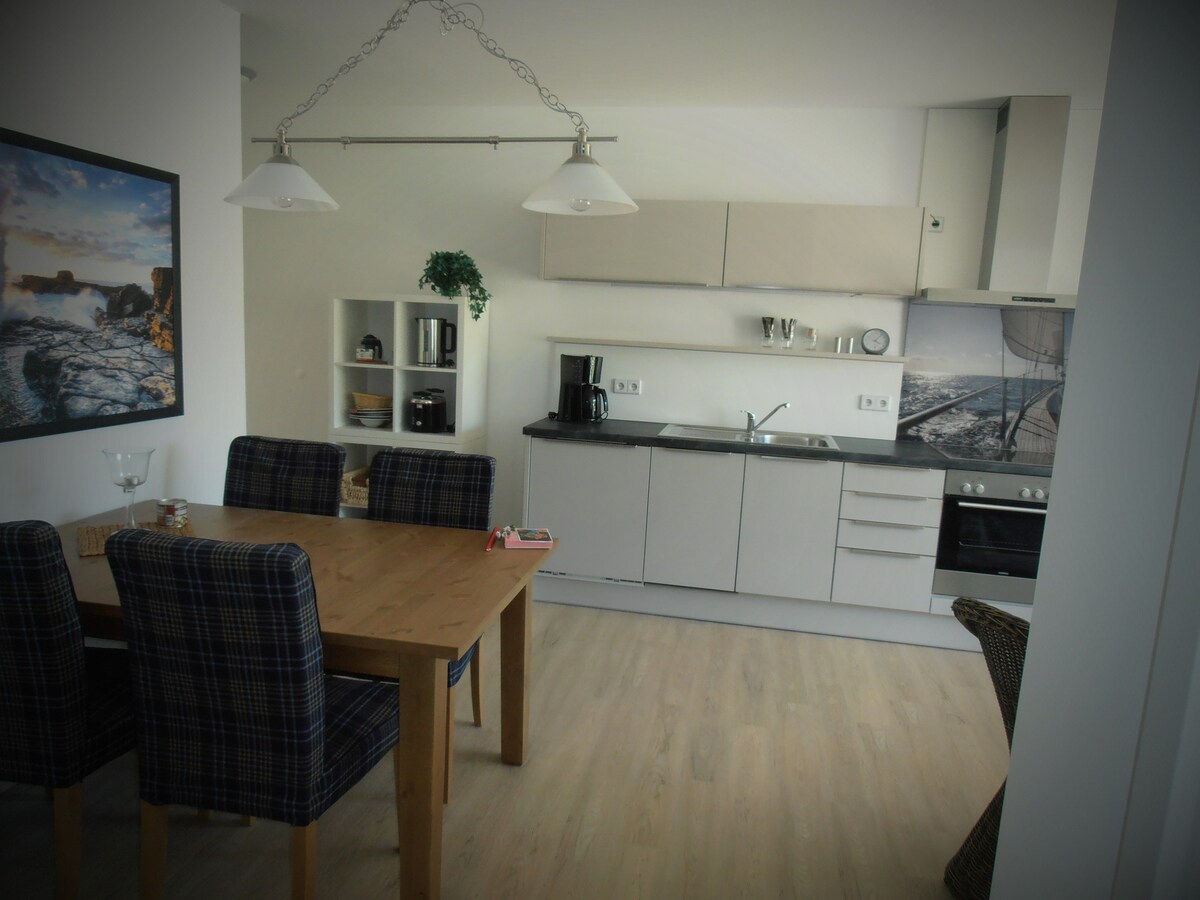 A modern kitchen area features a wooden dining table accompanied by four patterned chairs. Sleek cabinetry lines the walls, with a coffee maker and kitchen appliances visible. Natural light illuminates the space, enhancing the clean, minimalist design.