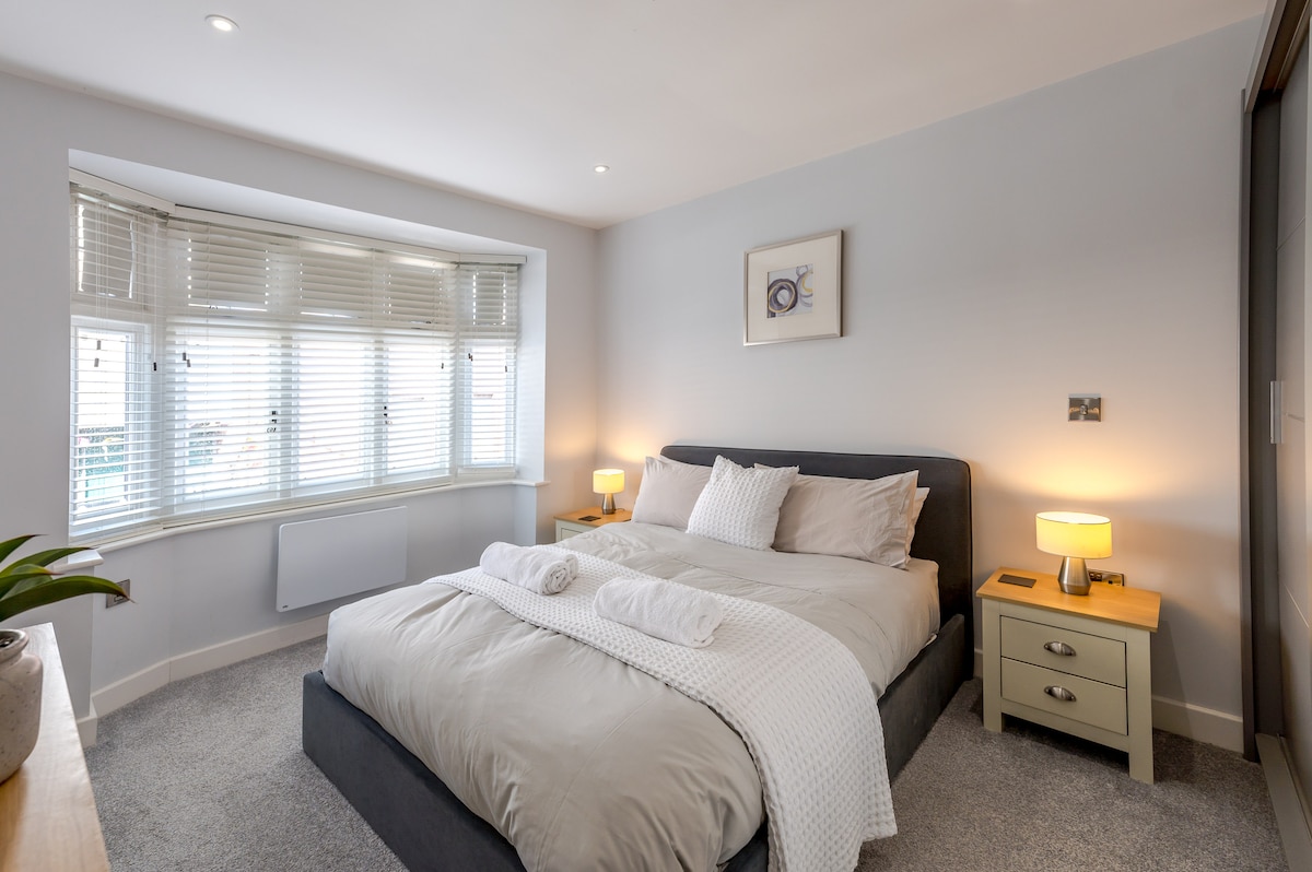 ห้องนอนที่สว่างและเงียบสงบพร้อมเตียงคู่ที่สะดวกสบาย ผ้าปูที่นอนสีขาวสดใส โต๊ะข้างเตียงพร้อมโคมไฟสีอบอุ่น พรมนอนุ่ม และหน้าต่างบานใหญ่พร้อมม่านที่ให้แสงธรรมชาติส่องเข้ามา