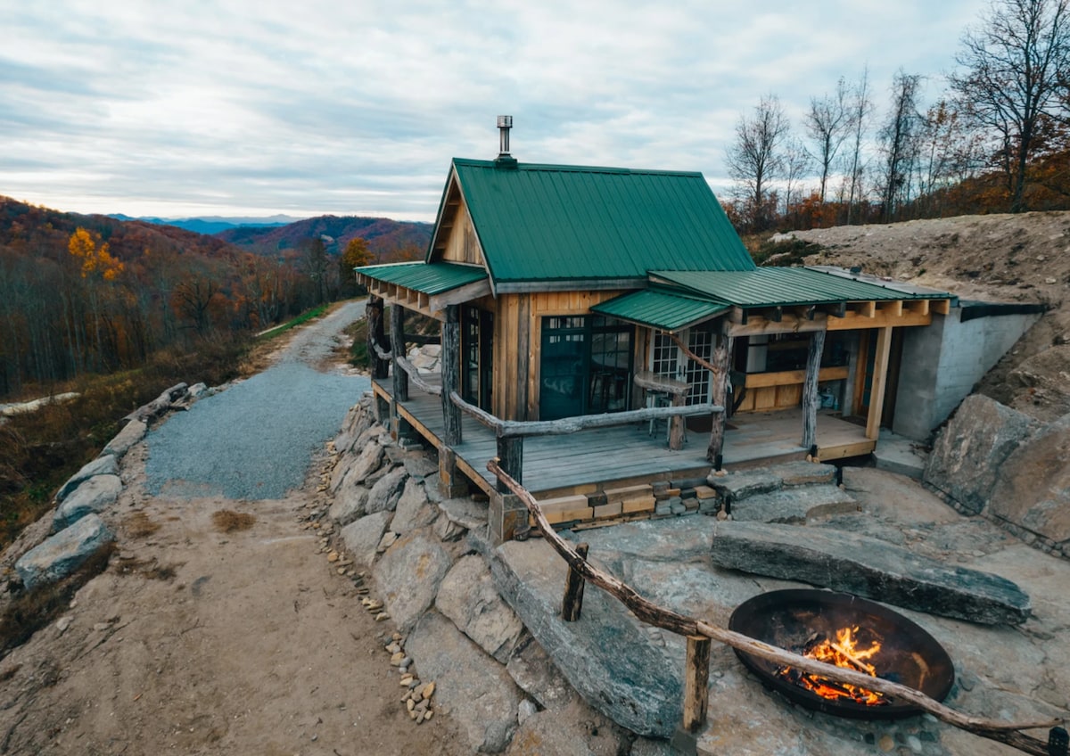 A rustic cottage is nestled among rocks, featuring a green metal roof and wooden beams. A spacious deck surrounds the cottage, with an outdoor fire pit visible in front. Scenic mountain landscapes extend in the background, showcasing a serene and natural setting.