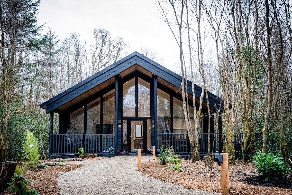 The lodge features a distinctive A-frame design with expansive glass panels that invite natural light. A gravel pathway leads to the entrance, framed by lush landscaping and surrounded by mature trees, creating a sense of seclusion within the woodland.