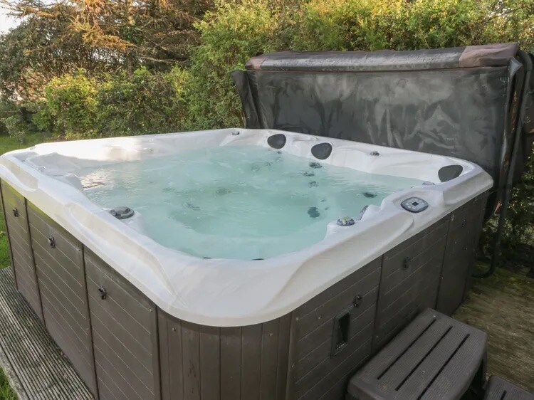 A large hot tub is situated outdoors, surrounded by lush greenery. The tub features multiple jets and a sturdy cover, with steps leading up for easy access. The tranquil water surface reflects the natural surroundings, creating a peaceful retreat for guests.