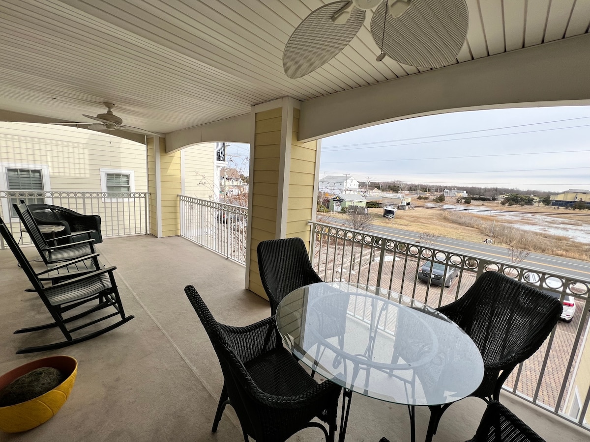 A spacious balcony is featured, equipped with a round glass dining table surrounded by four chairs. Two rocking chairs provide additional seating. The view showcases a serene landscape, with open space and distant buildings visible beyond the railing.