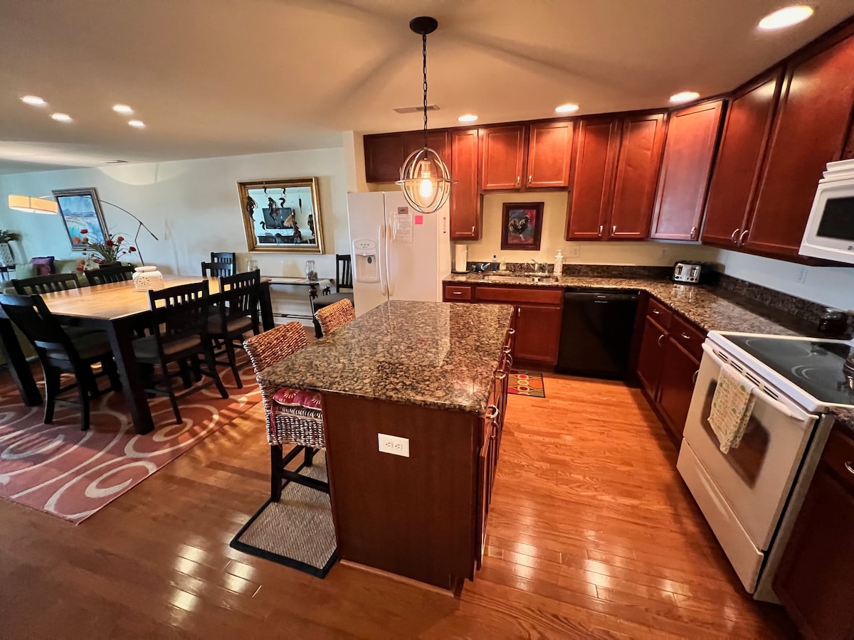 A modern kitchen features dark wooden cabinetry and granite countertops. An island with seating is positioned in the center, while a white stove and microwave are visible. The adjacent dining area showcases a large wooden table surrounded by black chairs, illuminated by warm overhead lighting.