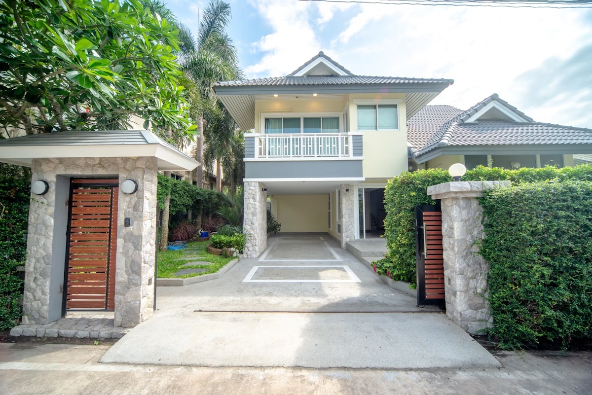A modern house is set amidst greenery, featuring a stone entrance with wooden gates. The driveway leads to an elevated porch with large glass doors, allowing natural light to illuminate the interior spaces. Lush plants and well-kept landscaping enhance the inviting exterior.